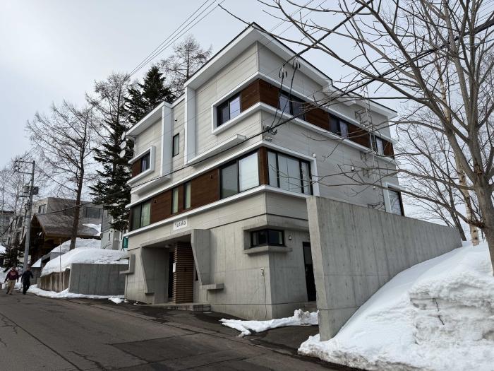Exterior view of modern Yutaka Townhouses in winter featuring contemporary architecture with wooden accents, concrete elements, multiple levels, and balconies, surrounded by snow-covered landscape in central Niseko, just minutes from Hirafu ski lifts.