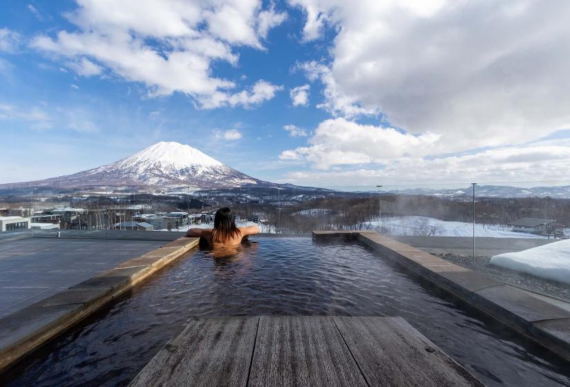 Enjoy the ultimate luxury of the Vale's 3 bedroom Yotei Panorama suite. A person sitting in an pool with Mount Yotei in the background.