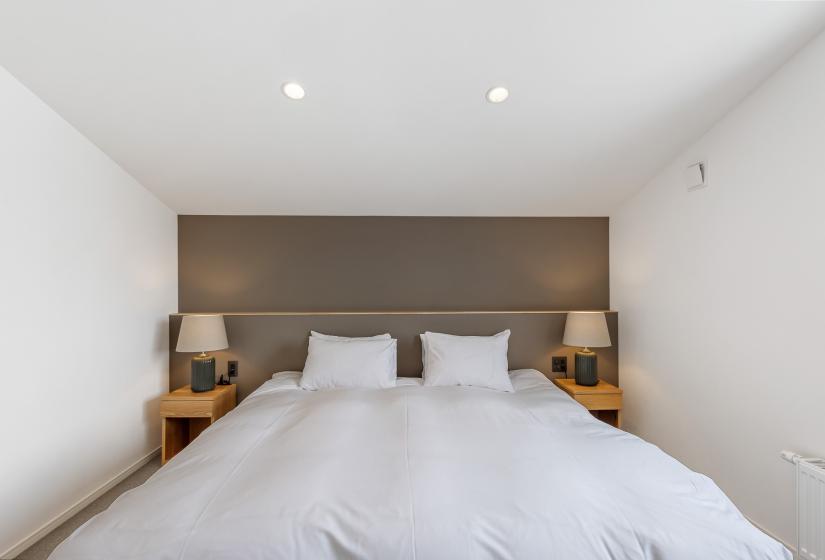 Minimalist bedroom featuring a king-size bed with white linens, built-in wooden bedside tables, and a contemporary grey feature wall headboard. Clean white walls and recessed lighting create a peaceful sleeping space
