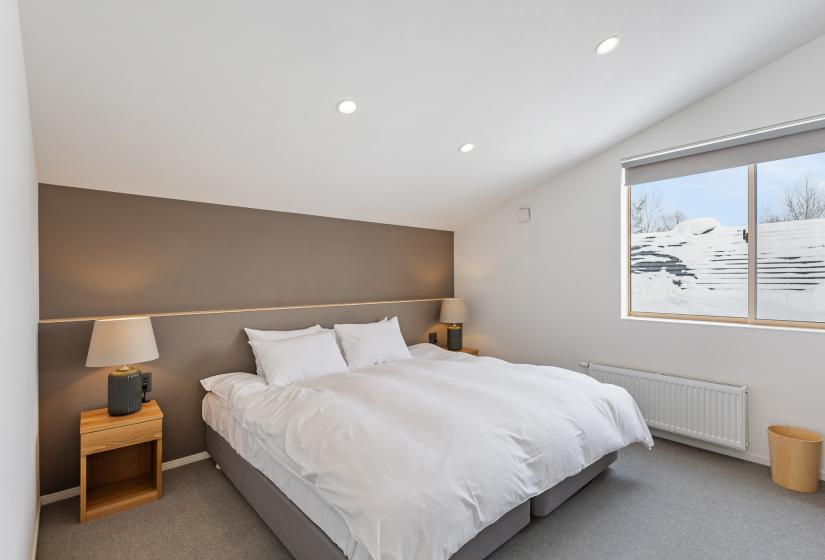 Bright guest bedroom with two twin beds featuring white linens, taupe accent wall, wooden bedside table with lamp, and a window offering natural light. Grey carpet adds warmth to the minimalist design