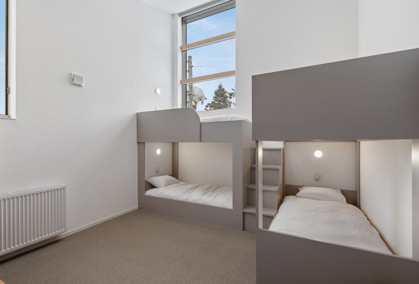 Modern bunk room featuring two sets of custom-built bunk beds with grey accent panels and white bedding. High windows provide natural light, and a wall-mounted radiator ensures comfort in the minimalist space.