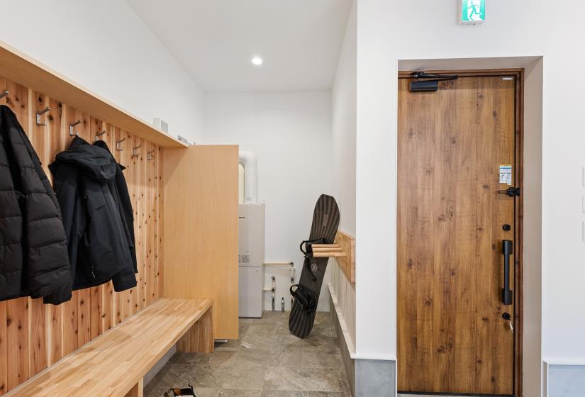 Dedicated ski room with wooden paneled walls featuring coat hooks, a built-in wooden bench for seating, and concrete flooring. The room includes winter gear storage and a wooden entry door, with an emergency exit sign mounted above