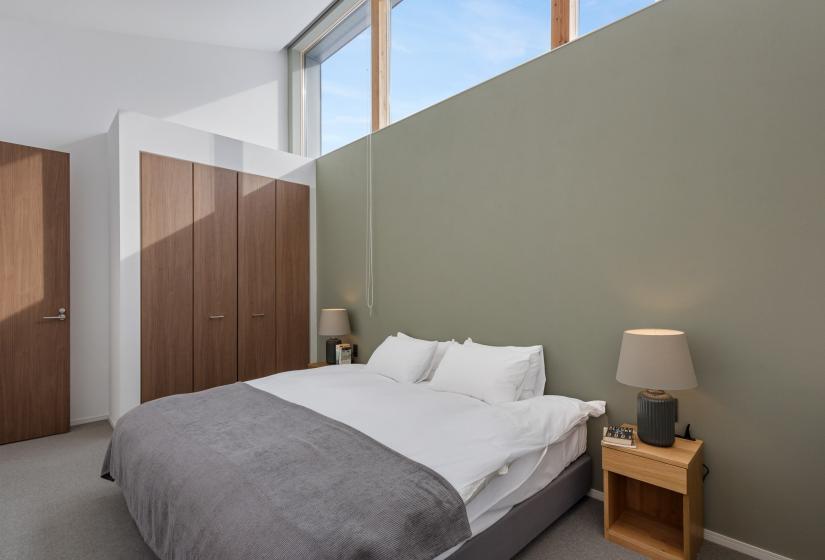 Master bedroom featuring a double bed with white linens and grey throw, set against a sage green accent wall. Built-in wooden wardrobes and high windows create a serene, contemporary atmosphere