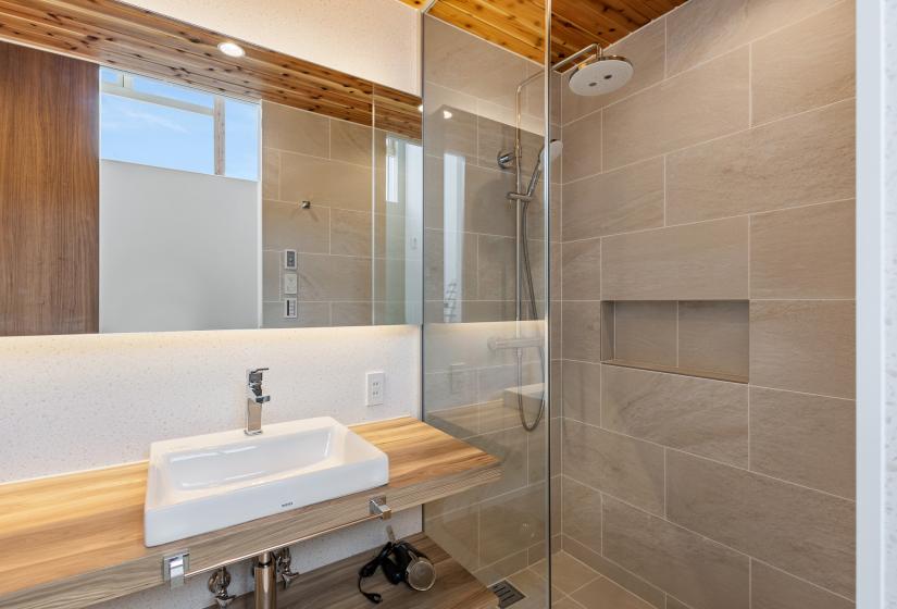 Modern ensuite bathroom featuring grey tile walls, glass shower enclosure, floating wooden vanity with white sink, and a wide mirror. Natural wood ceiling adds warmth to the contemporary design