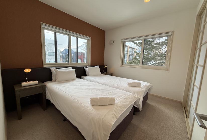 Stylish twin bedroom at Yutaka Townhouses with two comfortable beds dressed in white linens, bedside tables with reading lamps, large windows overlooking snowy Niseko forest, and warm wood accent wall creating a cozy mountain retreat atmosphere.